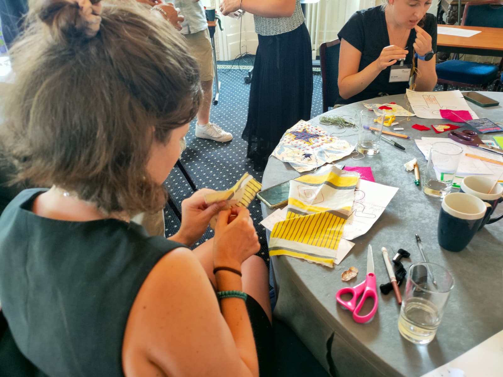A student with shoulder length brown hair sits with her back to the camera, working at a table. In her hands is a piece of colourful fabric, which she appears to be embroidering.